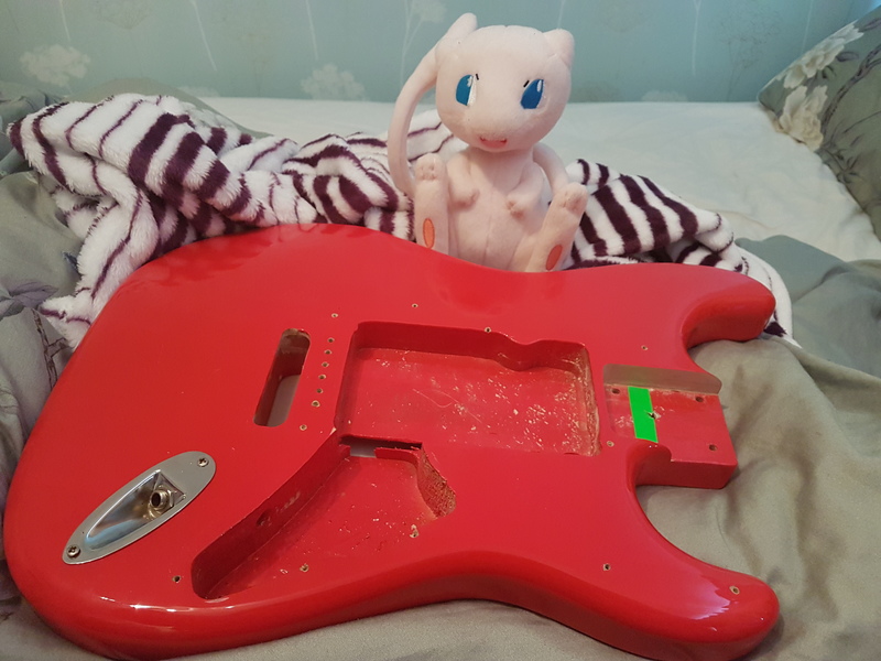 A red guitar body on a bed, in front of a Mew plush toy.